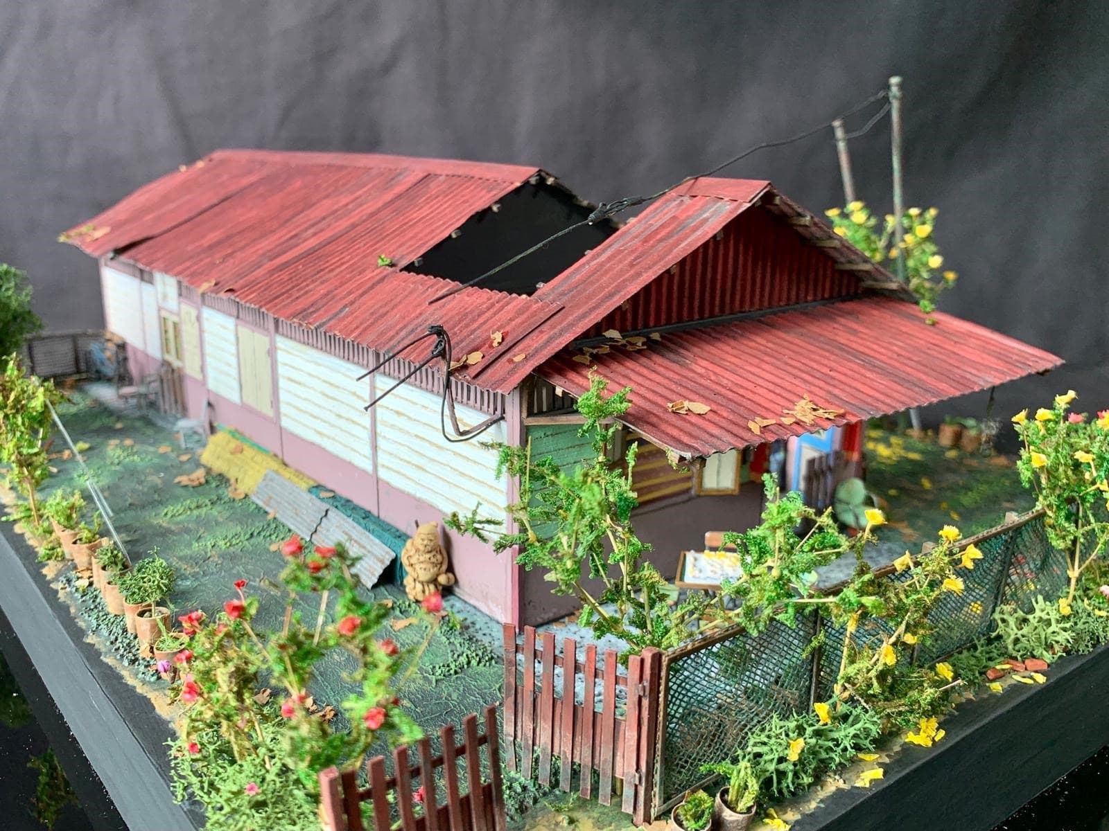 A detailed scale model of a traditional kampong house featuring a red corrugated zinc roof, wooden siding, and a small garden with potted plants and a picket fence.