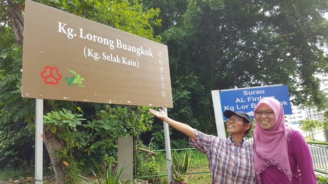 Two women posing and pointing towards a brown wooden sign for "Kg. Lorong Buangkok (Kg. Selak Kain)" with a lush green forest background.