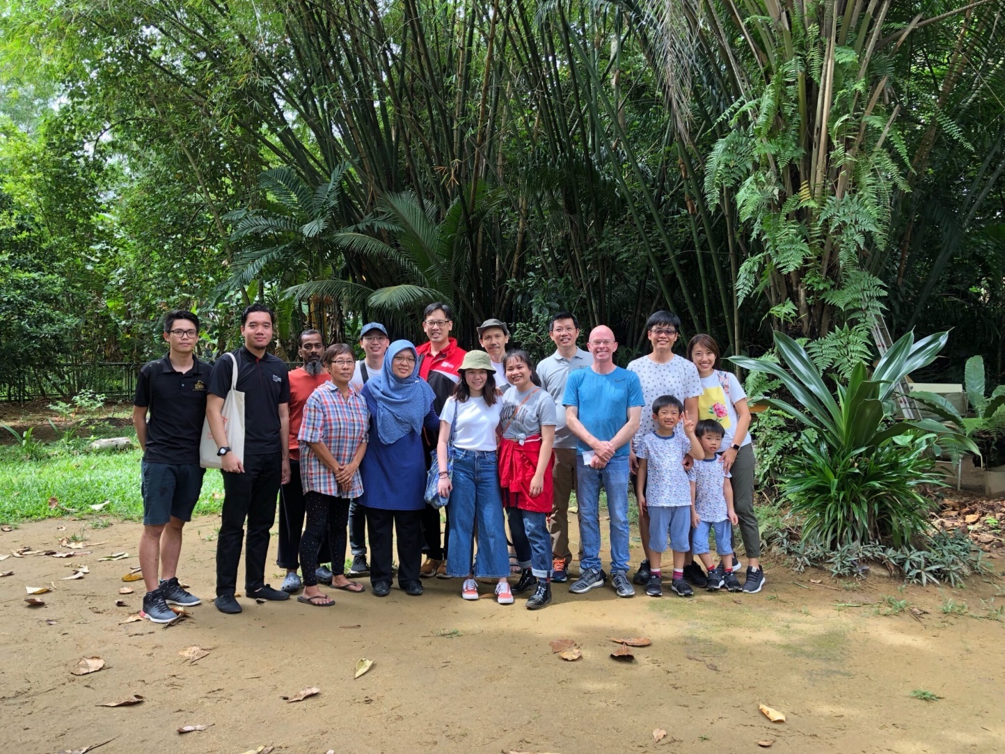A group of approximately fifteen people of diverse ages and backgrounds posing for a photo in front of a dense bamboo and tropical plant grove.