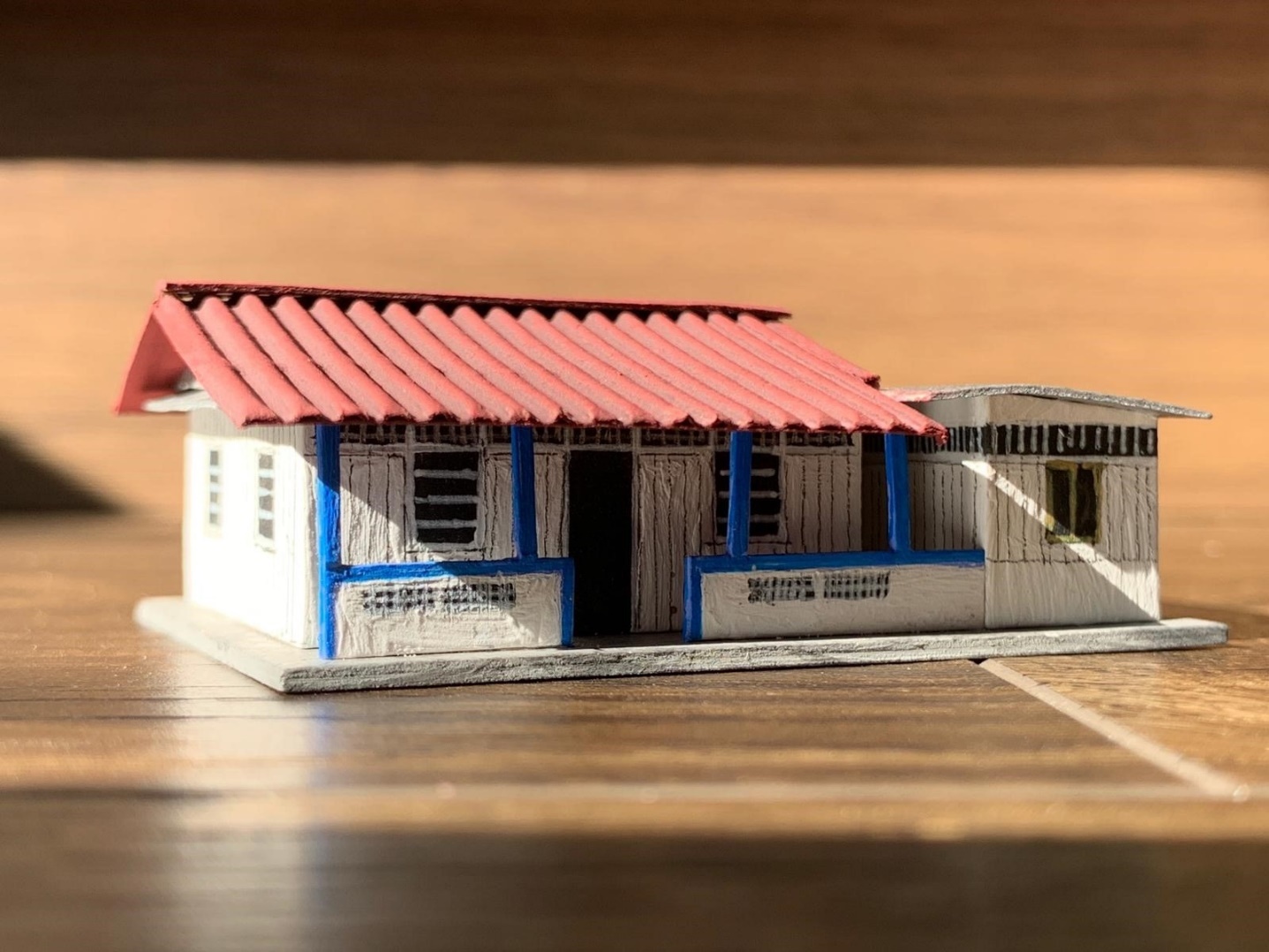 A simple architectural scale model of a white kampong house with a red roof and blue trim, shown in close-up on a wooden surface.