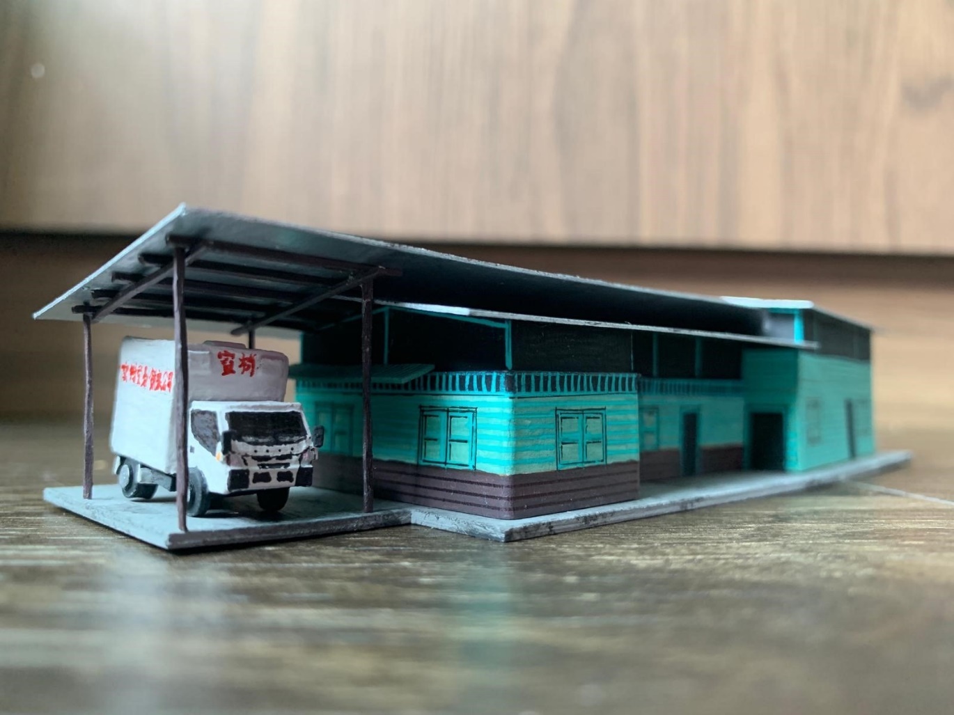 A scale model of a long teal-colored building with a large grey overhang, featuring a small white delivery truck parked underneath the shelter.