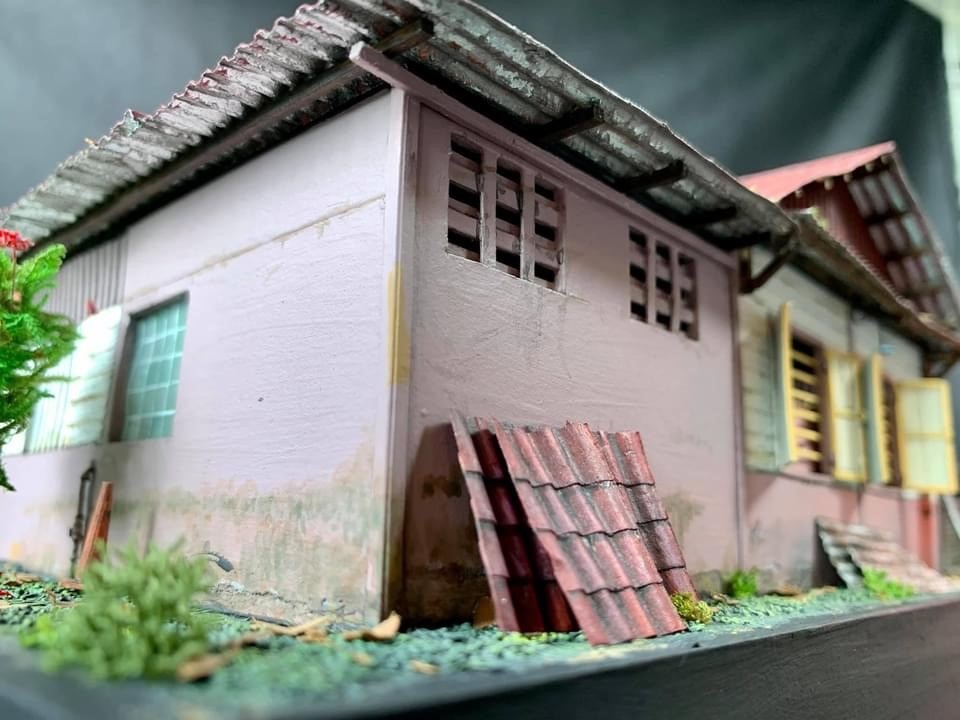 A low-angle close-up of a pink kampong house model, showing realistic weathering on the walls and pieces of corrugated roofing leaning against the side.