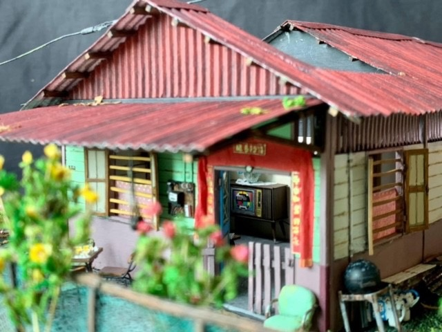 A detailed close-up of a green kampong house model’s entrance, showing red banners over the door and a tiny vintage television visible inside.