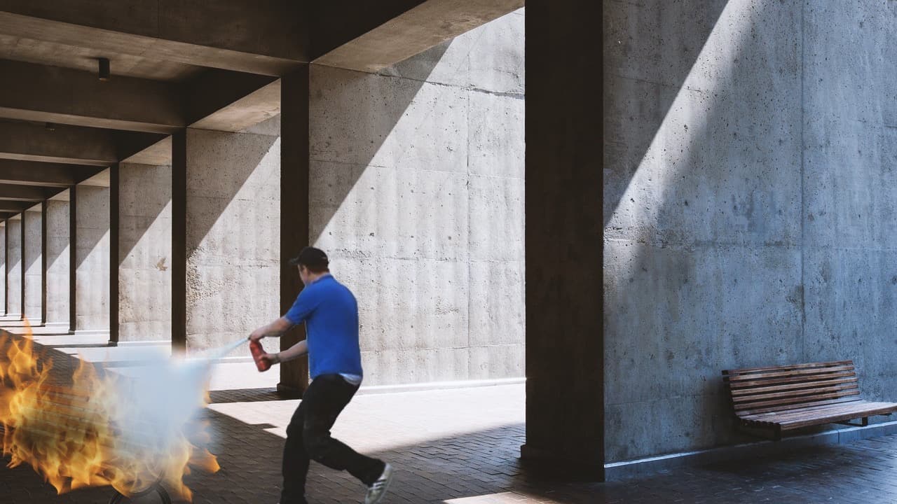 A man in a blue shirt uses a red fire extinguisher to put out a fire on a wooden bench in a concrete architectural hallway.
