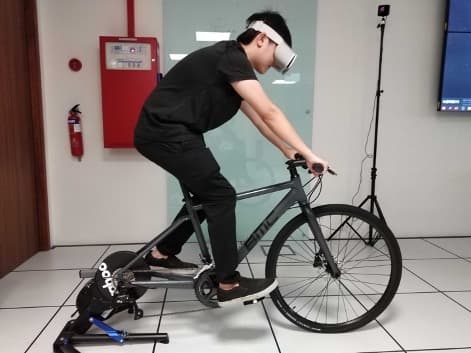 A person wearing a VR headset while riding a grey bicycle mounted on a Wahoo smart indoor trainer in a laboratory setting. A motion tracking sensor on a tripod is visible in the background.