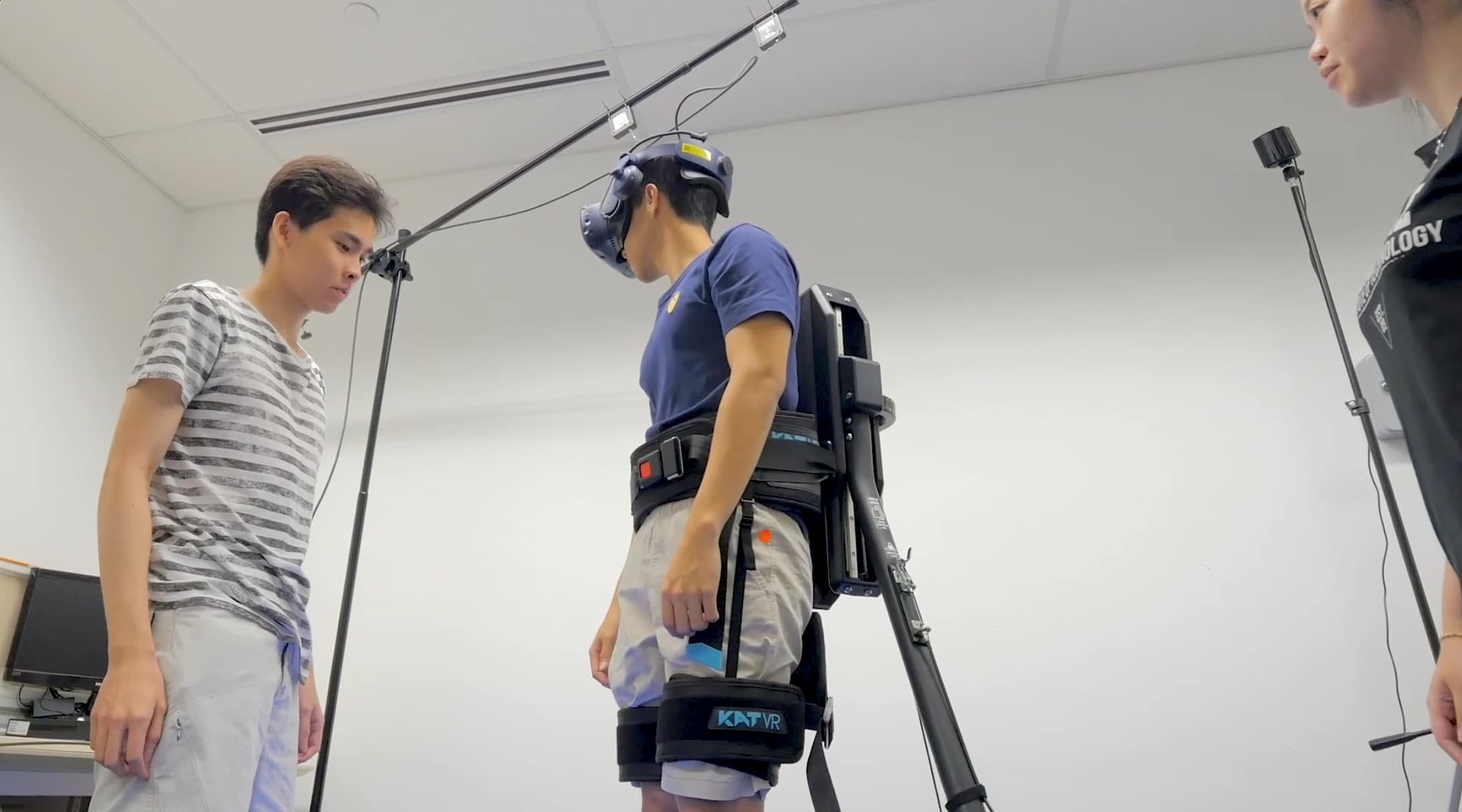 A person wearing a VR headset and a KAT VR haptic body suit and harness system, standing in a laboratory while two researchers observe. The setup includes overhead sensors and motion tracking equipment for an immersive virtual reality simulation.