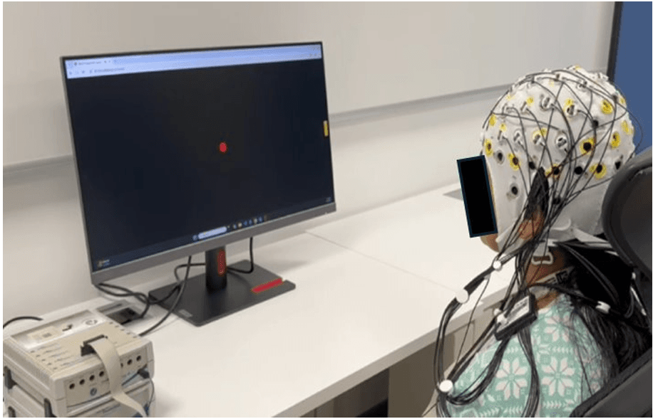 A person participating in an EEG experiment, wearing a sensor-laden cap connected to a recording device, while watching a computer monitor displaying a red dot.