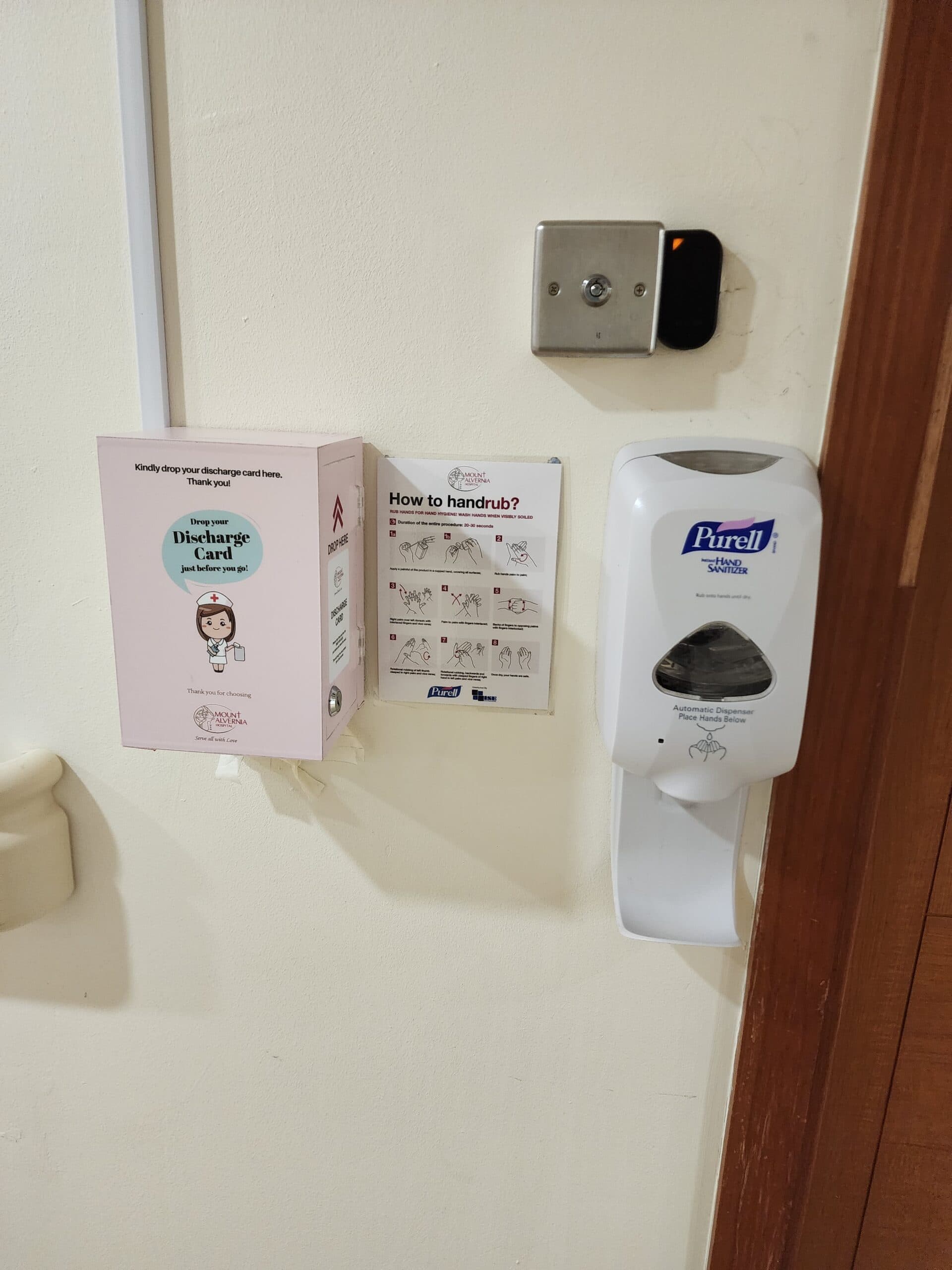 A wall-mounted hospital station featuring a pink box for "Discharge Card" collection, an instructional sign titled "How to handrub?" with diagrams on proper sanitization techniques, and an automatic Purell hand sanitizer dispenser.